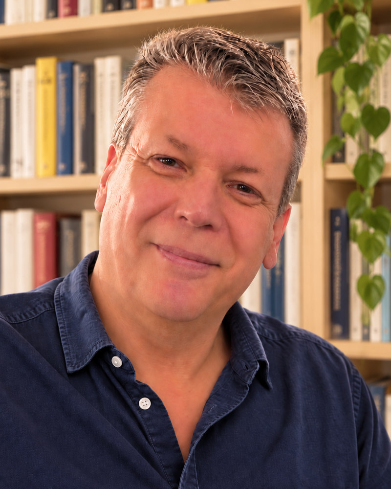 Glenn Carey smiling in front of bookshelves and a houseplant